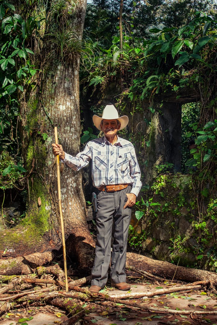 Elderly man in a white cowboy hat holding a stick amidst a lush green forest backdrop.