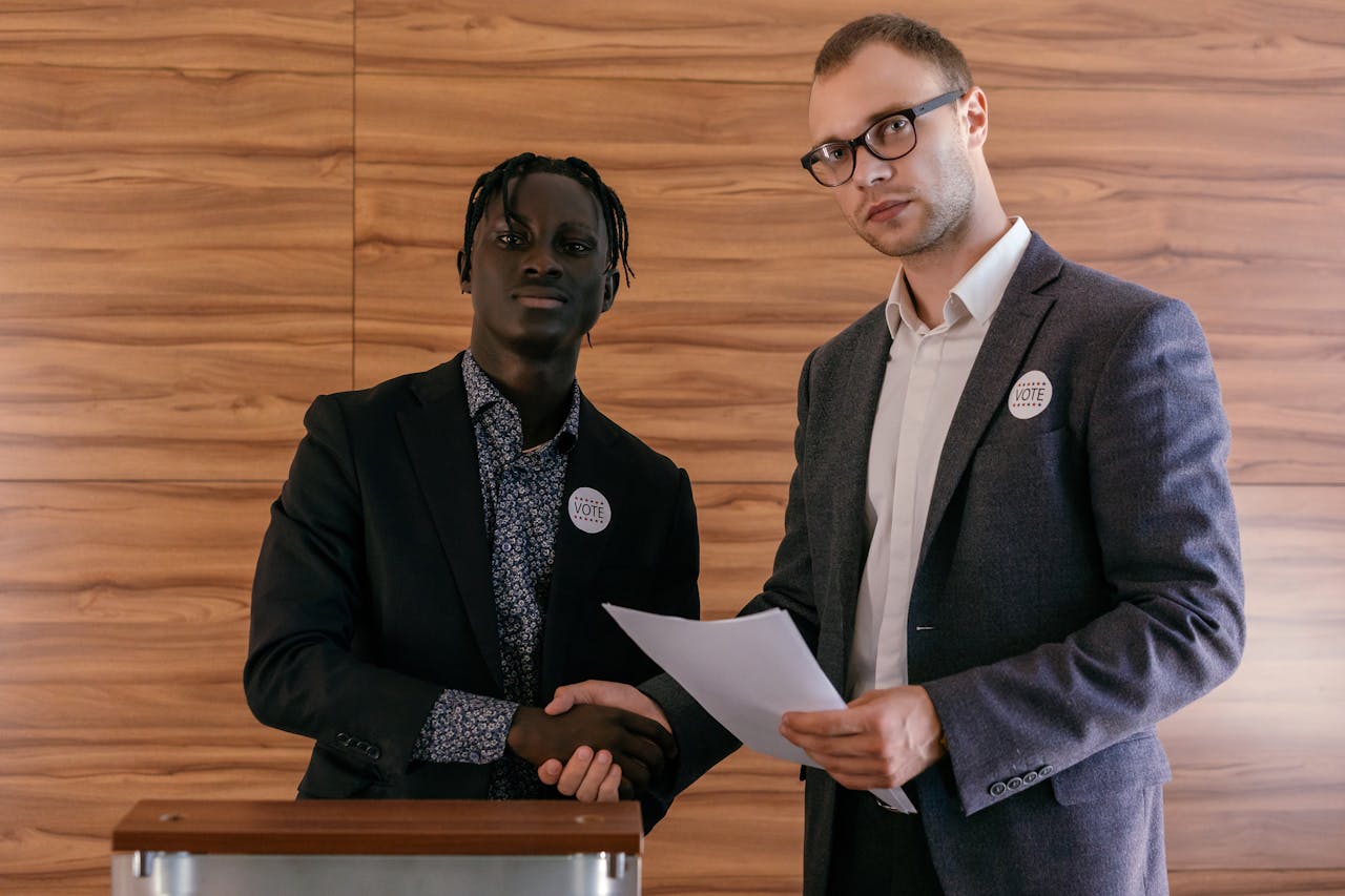 Two men in business attire shaking hands with agreement, promoting voting indoors.